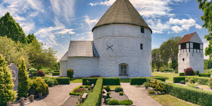 Bornholm_189566410 Rundkirche auf der Insel Bornholm, Dänemark, mit weiß verputzten Mauern, kegelförmigem Dach und gepflegtem Garten bei blauem Himmel.