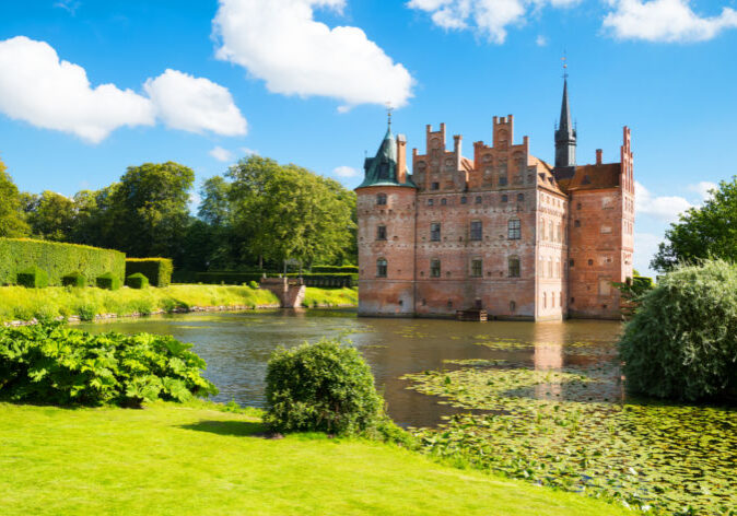 Busreisen nach Daenemark. Schloss Egeskov auf der Insel Fünen, Dänemark, an einem Sommertag mit Wassergraben, grüner Parkanlage und blauem Himmel.