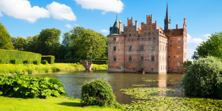 Busreisen nach Daenemark. Schloss Egeskov auf der Insel Fünen, Dänemark, an einem Sommertag mit Wassergraben, grüner Parkanlage und blauem Himmel.