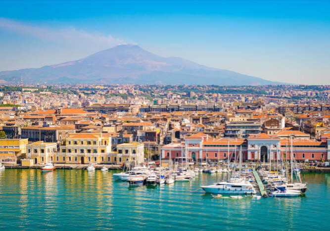 Catania_165729677 Hafen von Catania auf Sizilien, Italien, mit Segelbooten, historischen Gebäuden und dem Ätna im Hintergrund.