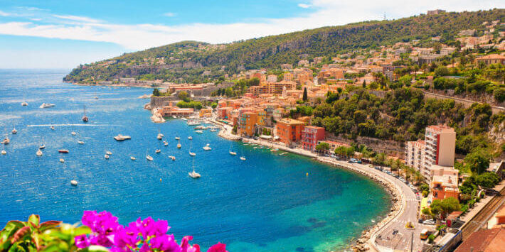 Cote dAzur Frankreich Reisen. Küstenblick auf die Côte d’Azur, Frankreich, mit türkisblauem Meer, Booten, bunten Häusern und blühenden Bougainvilleen im Vordergrund.