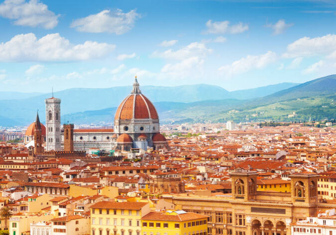 Blick über Florenz, Italien, mit der Kathedrale Santa Maria del Fiore und ihrer markanten roten Kuppel, dem Glockenturm von Giotto und den umliegenden Dächern vor einer Bergkulisse.