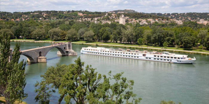 Flusskreuzfahrtschiff auf der Rhône in Avignon, Frankreich, mit Blick auf die Pont Saint-Bénézet und grüne Uferlandschaft.