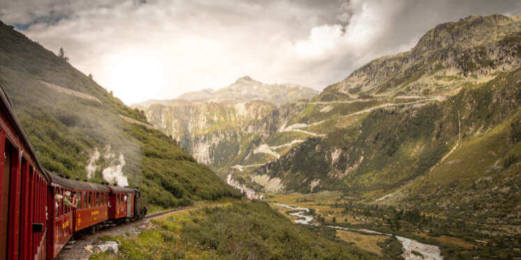 Historische Furka-Dampfbahn fährt durch das Gebirge ins Rhonetal, Wallis, Schweiz, mit Flusslauf und Bergkulisse im Hintergrund.