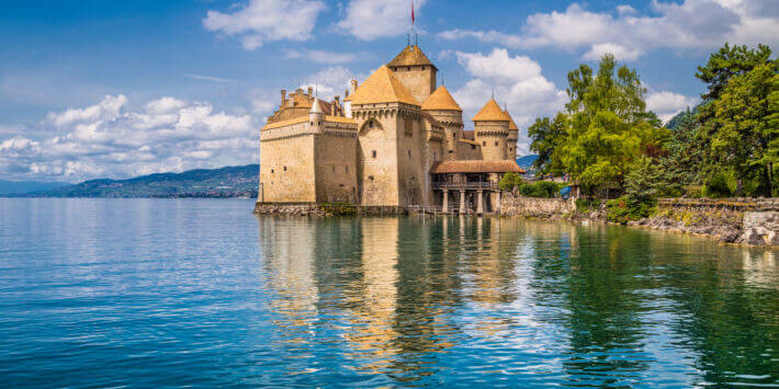 Reise Inspirationen Schweiz Reisen mit Blick auf Genf. Schloss Chillon am Ufer des Genfersees, Schweiz, mit Spiegelung im Wasser und Alpenpanorama im Hintergrund.