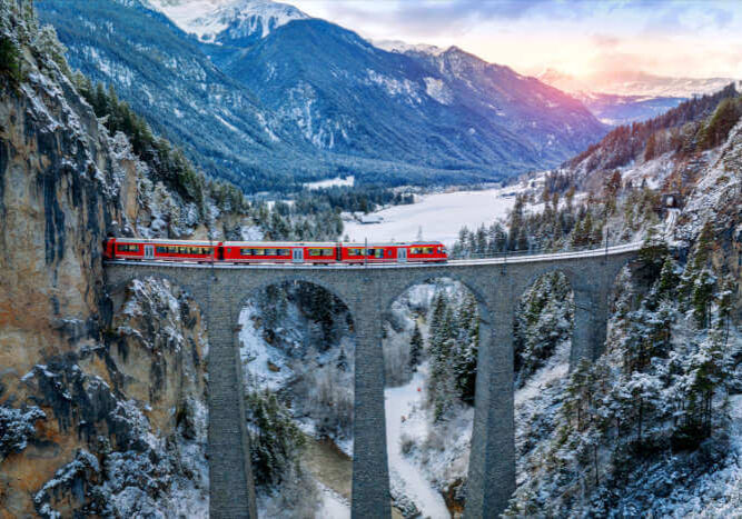 Roter Glacier-Express auf dem Landwasserviadukt in der verschneiten Winterlandschaft der Schweizer Alpen bei Sonnenuntergang.