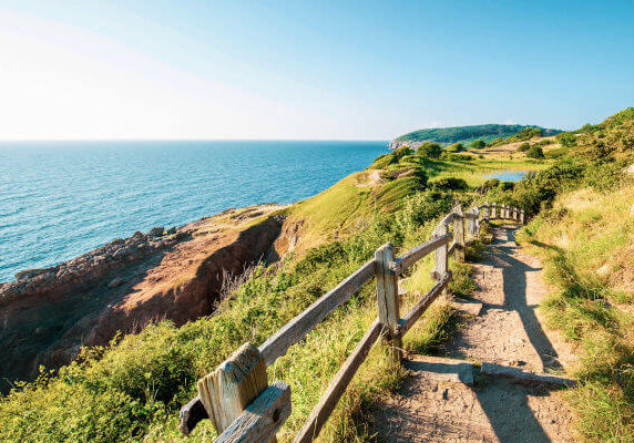 Busreisen nach Bornholm. Küstenwanderweg auf der Insel Bornholm, Dänemark, mit Holzgeländer, grünen Wiesen, Felsen und Blick auf das blaue Meer.