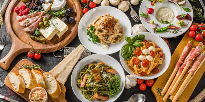 Italien_189829853 Auswahl italienischer Spezialitäten mit Pasta, Käse, Antipasti, Brot und Schinken auf rustikalem Holztisch.