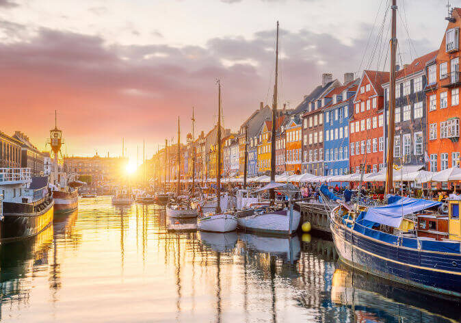 Busreisen nach Kopenhagen. Sonnenuntergang am Nyhavn in Kopenhagen, Dänemark, mit bunten Häuserfassaden, Segelbooten im Hafen und ruhigem Wasser.