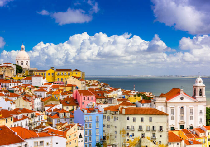 Staedtereisen nach Lissabon. Bunte Häuserfassaden im Stadtteil Alfama in Lissabon, Portugal, mit Blick auf den Tejo unter blauem Himmel mit Wolken.