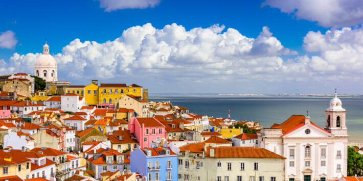 Staedtereisen nach Lissabon. Bunte Häuserfassaden im Stadtteil Alfama in Lissabon, Portugal, mit Blick auf den Tejo unter blauem Himmel mit Wolken.